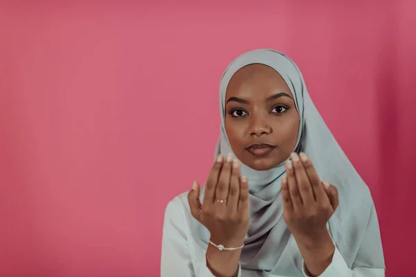 Modern afrikansk Muselman kvinna gör traditionell bön till Gud, håller händerna i bedjande gest, bär traditionella vita kläder, har allvarliga ansiktsuttryck, isolerade över plast rosa bakgrund — Stockfoto