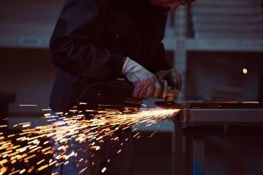 Heavy Industry Engineering Factory Interior with Industrial Worker Using Angle Grinder and Cutting a Metal Tube. Güvenlik Üniforması ve Sert Şapka Üretim Metal Yapıları Müteahhiti.