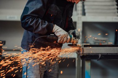 Heavy Industry Engineering Factory Interior with Industrial Worker Using Angle Grinder and Cutting a Metal Tube. Güvenlik Üniforması ve Sert Şapka Üretim Metal Yapıları Müteahhiti.