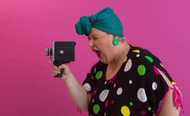 Artı, komik duygusal yüz ifadesi olan, pembe arka planda izole edilmiş eski moda kameralı, yaz modası tarzında gezgin, heyecanlı turist tipli, gülen bir kadın..