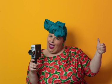 Artı, komik duygusal yüz ifadesi olan, sarı arka planda izole edilmiş eski moda kameralı, yaz modası tarzında gezgin, heyecanlı turist tipli, güler yüzlü bir kadın..