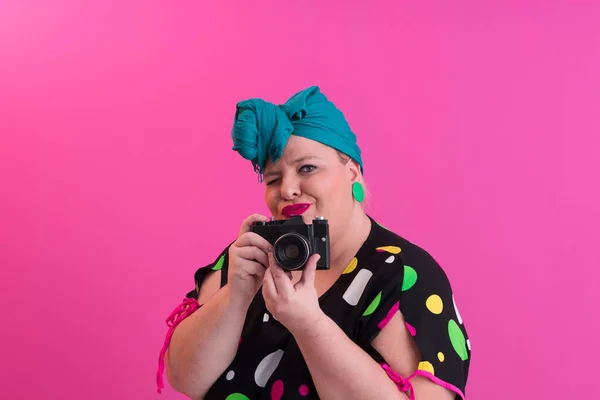 Plus size lachende vrouw met grappige emotionele gezicht uitdrukking met vintage camera in denim jurk geïsoleerd op roze achtergrond, reiziger op vakantie, zomer mode stijl, opgewonden toerist. — Stockfoto