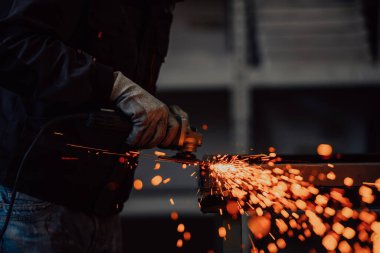 Heavy Industry Engineering Factory Interior with Industrial Worker Using Angle Grinder and Cutting a Metal Tube. Güvenlik Üniforması ve Sert Şapka Üretim Metal Yapıları Müteahhiti.