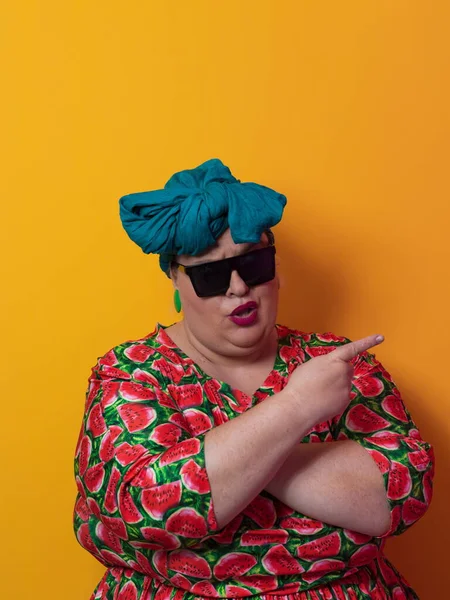 Funny plus size sexy model woman with trendy sunglasses, fashionable overweight blonde in colorful dress posing at the yellow background in studio. — Zdjęcie stockowe