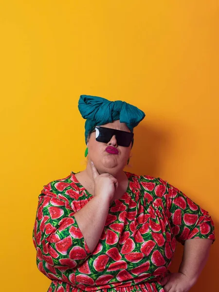 Komik artı boyut seksi, güneş gözlüklü, şık, şişman, renkli elbiseli, stüdyoda sarı arka planda poz veren bir kadın..