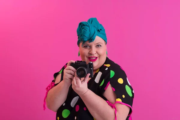 Plus size smiling woman with funny emotional face expression with vintage camera in denim dress isolated on pink background, traveler on vacation, summer fashion style, excited tourist. — Stock Photo, Image