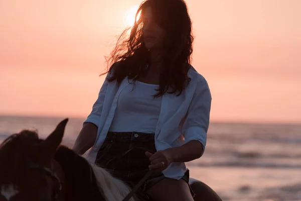 Woman in summer clothes enjoys riding a horse on a beautiful sandy beach at sunset. Selective focus — Stock Fotó