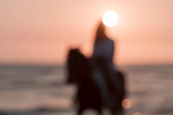 Ett suddigt foto. Kvinnan i sommarkläder njuter av att rida på en häst på en vacker sandstrand vid solnedgången. Selektiv inriktning — Stockfoto