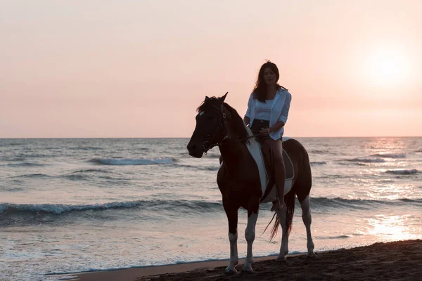 Mujer en ropa de verano disfruta montar a caballo en una hermosa playa de arena al atardecer. Enfoque selectivo — Foto de Stock