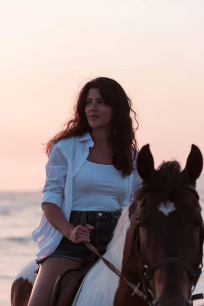 Woman in summer clothes enjoys riding a horse on a beautiful sandy beach at sunset. Selective focus