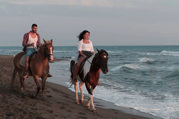 A loving young couple in summer clothes riding a horse on a sandy beach at sunset. Sea and sunset in the background. Selective focus —  Fotos de Stock