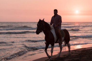 Yaz kıyafetleri içinde modern bir adam gün batımında kumlu güzel bir sahilde ata binmekten hoşlanır. Seçici odak 