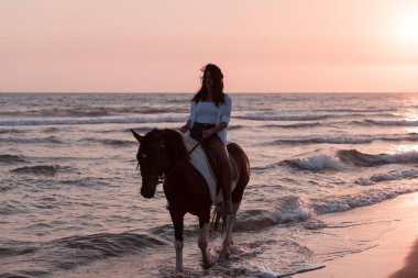 Yaz kıyafetleri içinde bir kadın gün batımında kumlu güzel bir sahilde ata binmekten hoşlanır. Seçici odak 