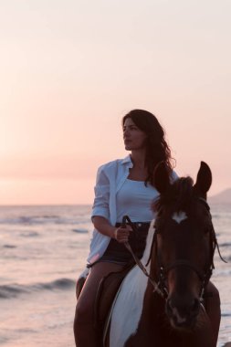 Yaz kıyafetleri içinde bir kadın gün batımında kumlu güzel bir sahilde ata binmekten hoşlanır. Seçici odak 