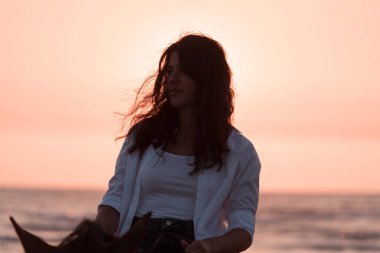 Yaz kıyafetleri içinde bir kadın gün batımında kumlu güzel bir sahilde ata binmekten hoşlanır. Seçici odak 