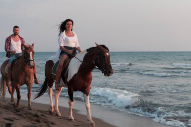 Gün batımında kumlu bir sahilde ata binen yaz kıyafetleri içinde sevgi dolu bir çift. Arka planda deniz ve gün batımı. Seçici odak 