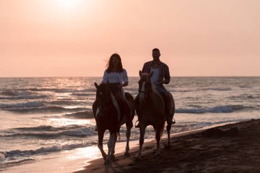 Gün batımında kumlu bir sahilde ata binen yaz kıyafetleri içinde sevgi dolu bir çift. Arka planda deniz ve gün batımı. Seçici odak 