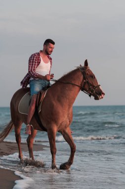 Yaz kıyafetleri içinde modern bir adam gün batımında kumlu güzel bir sahilde ata binmekten hoşlanır. Seçici odak 