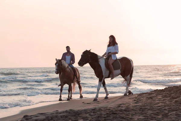Ett kärleksfullt par i sommarkläder ridande på en häst på en sandstrand vid solnedgången. Hav och solnedgång i bakgrunden. Selektiv inriktning — Stockfoto