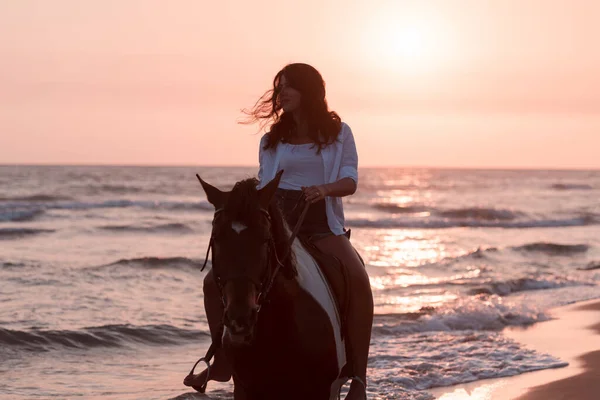 Woman in summer clothes enjoys riding a horse on a beautiful sandy beach at sunset. Selective focus — Stockfoto