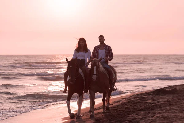 Um casal amoroso em roupas de verão montando um cavalo em uma praia de areia ao pôr do sol. Mar e pôr do sol no fundo. Foco seletivo — Fotografia de Stock