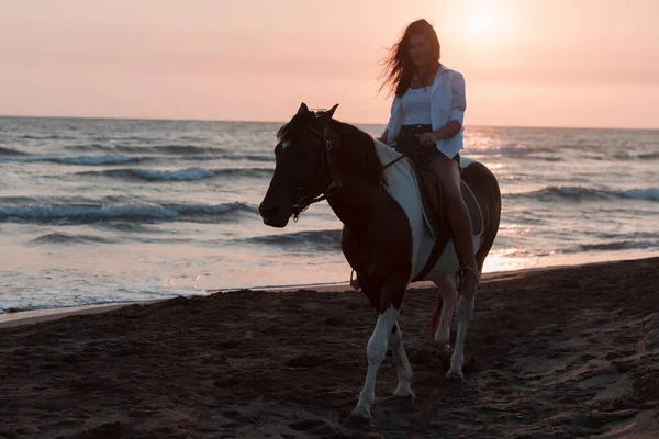 Γυναίκα με καλοκαιρινά ρούχα απολαμβάνει ιππασία σε μια όμορφη αμμώδη παραλία το ηλιοβασίλεμα. Επιλεκτική εστίαση — Φωτογραφία Αρχείου