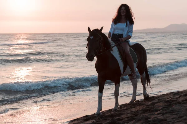 Woman in summer clothes enjoys riding a horse on a beautiful sandy beach at sunset. Selective focus