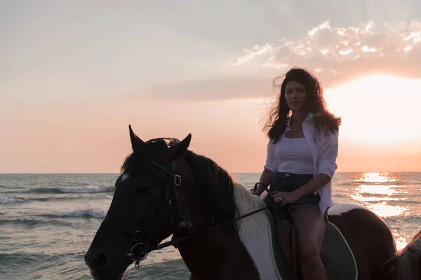 Woman in summer clothes enjoys riding a horse on a beautiful sandy beach at sunset. Selective focus — Zdjęcie stockowe