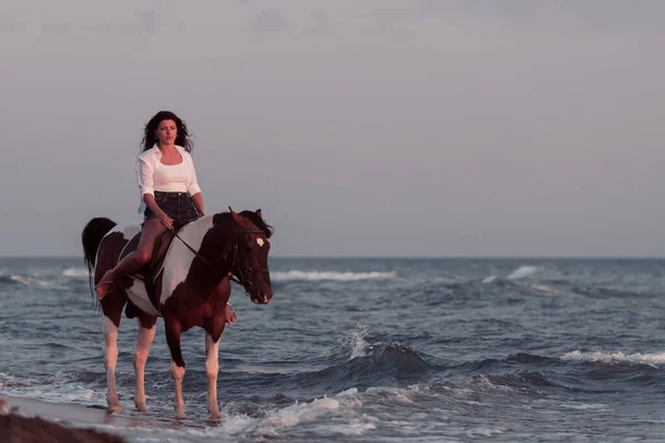 Woman in summer clothes enjoys riding a horse on a beautiful sandy beach at sunset. Selective focus