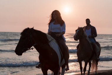 Gün batımında kumlu bir sahilde ata binen yaz kıyafetleri içinde sevgi dolu bir çift. Arka planda deniz ve gün batımı. Seçici odak 