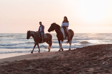 Gün batımında kumlu bir sahilde ata binen yaz kıyafetleri içinde sevgi dolu bir çift. Arka planda deniz ve gün batımı. Seçici odak 