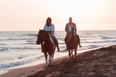 Gün batımında kumlu bir sahilde ata binen yaz kıyafetleri içinde sevgi dolu bir çift. Arka planda deniz ve gün batımı. Seçici odak 