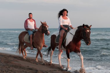 Gün batımında kumlu bir sahilde ata binen yaz kıyafetleri içinde sevgi dolu bir çift. Arka planda deniz ve gün batımı. Seçici odak 