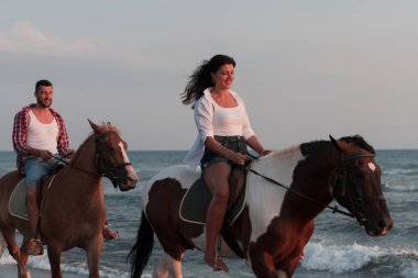 Gün batımında kumlu bir sahilde ata binen yaz kıyafetleri içinde sevgi dolu bir çift. Arka planda deniz ve gün batımı. Seçici odak 