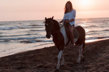 Yaz kıyafetleri içinde bir kadın gün batımında kumlu güzel bir sahilde ata binmekten hoşlanır. Seçici odak 