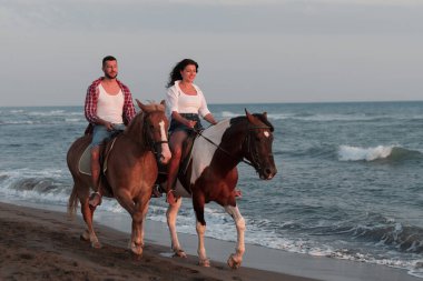 Gün batımında kumlu bir sahilde ata binen yaz kıyafetleri içinde sevgi dolu bir çift. Arka planda deniz ve gün batımı. Seçici odak 