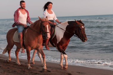 Gün batımında kumlu bir sahilde ata binen yaz kıyafetleri içinde sevgi dolu bir çift. Arka planda deniz ve gün batımı. Seçici odak 