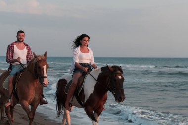 Gün batımında kumlu bir sahilde ata binen yaz kıyafetleri içinde sevgi dolu bir çift. Arka planda deniz ve gün batımı. Seçici odak 