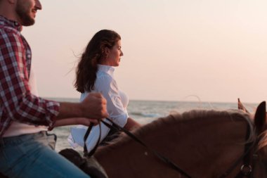 Gün batımında kumlu bir sahilde ata binen yaz kıyafetleri içinde sevgi dolu bir çift. Arka planda deniz ve gün batımı. Seçici odak 