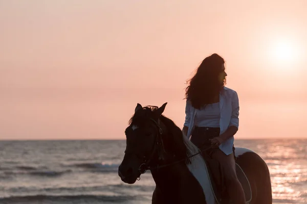 Mujer en ropa de verano disfruta montar a caballo en una hermosa playa de arena al atardecer. Enfoque selectivo —  Fotos de Stock