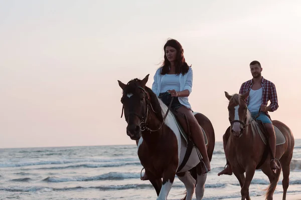 Ein liebes Paar in Sommerkleidung, das bei Sonnenuntergang auf einem Pferd an einem Sandstrand reitet. Meer und Sonnenuntergang im Hintergrund. Selektiver Fokus — Stockfoto