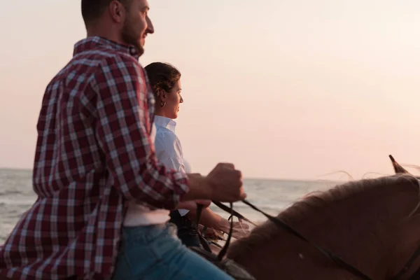 Una pareja amorosa en ropa de verano montando un caballo en una playa de arena al atardecer. Mar y puesta de sol en el fondo. Enfoque selectivo — Foto de Stock