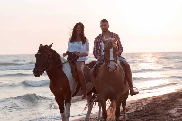 A loving couple in summer clothes riding a horse on a sandy beach at sunset. Sea and sunset in the background. Selective focus — Zdjęcie stockowe