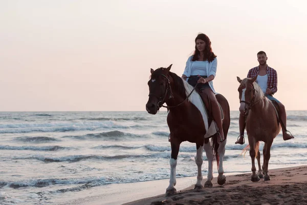 Una pareja amorosa en ropa de verano montando un caballo en una playa de arena al atardecer. Mar y puesta de sol en el fondo. Enfoque selectivo —  Fotos de Stock