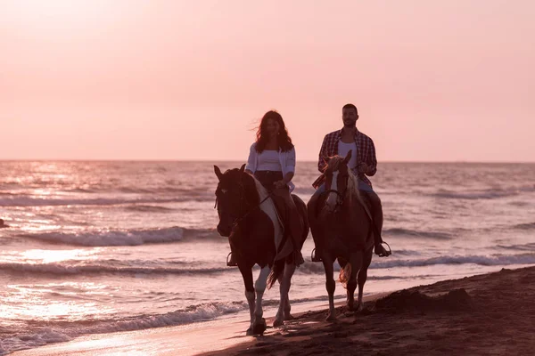 Um casal amoroso em roupas de verão montando um cavalo em uma praia de areia ao pôr do sol. Mar e pôr do sol no fundo. Foco seletivo — Fotografia de Stock