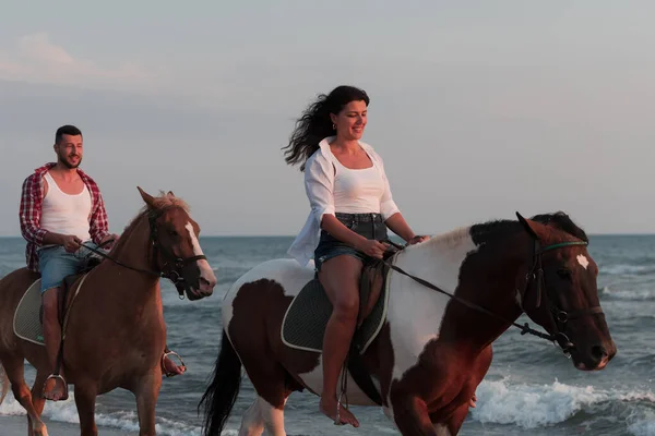 A loving couple in summer clothes riding a horse on a sandy beach at sunset. Sea and sunset in the background. Selective focus —  Fotos de Stock