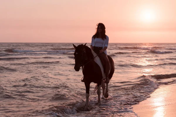 Woman in summer clothes enjoys riding a horse on a beautiful sandy beach at sunset. Selective focus — Fotografia de Stock