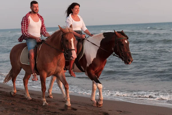 Gün batımında kumlu bir sahilde ata binen yaz kıyafetleri içinde sevgi dolu bir çift. Arka planda deniz ve gün batımı. Seçici odak 