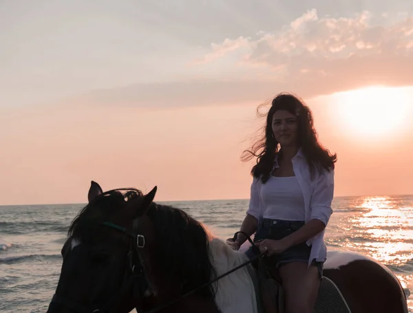 Mujer en ropa de verano disfruta montar a caballo en una hermosa playa de arena al atardecer. Enfoque selectivo —  Fotos de Stock