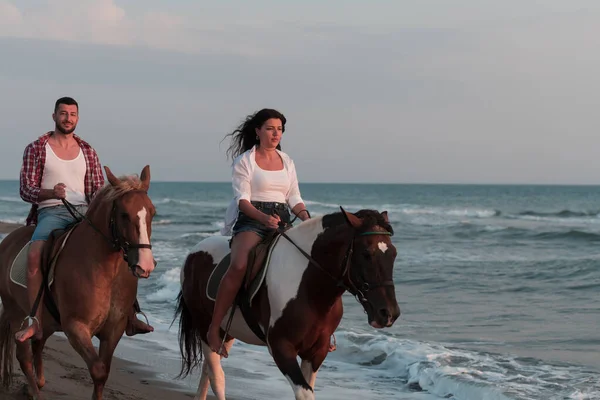 Una pareja amorosa en ropa de verano montando un caballo en una playa de arena al atardecer. Mar y puesta de sol en el fondo. Enfoque selectivo —  Fotos de Stock